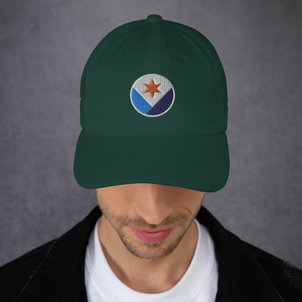 A man facing us wearing a spruce green Syracuse, NY hat with an embroidered Syracuse city flag design