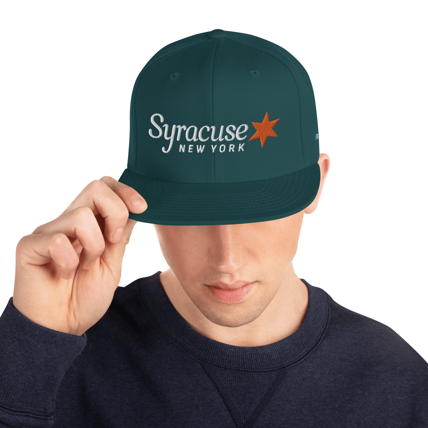 A man facing us wearing a spruce green Syracuse, NY city flag hat with an embroidered Syracuse, NY logo