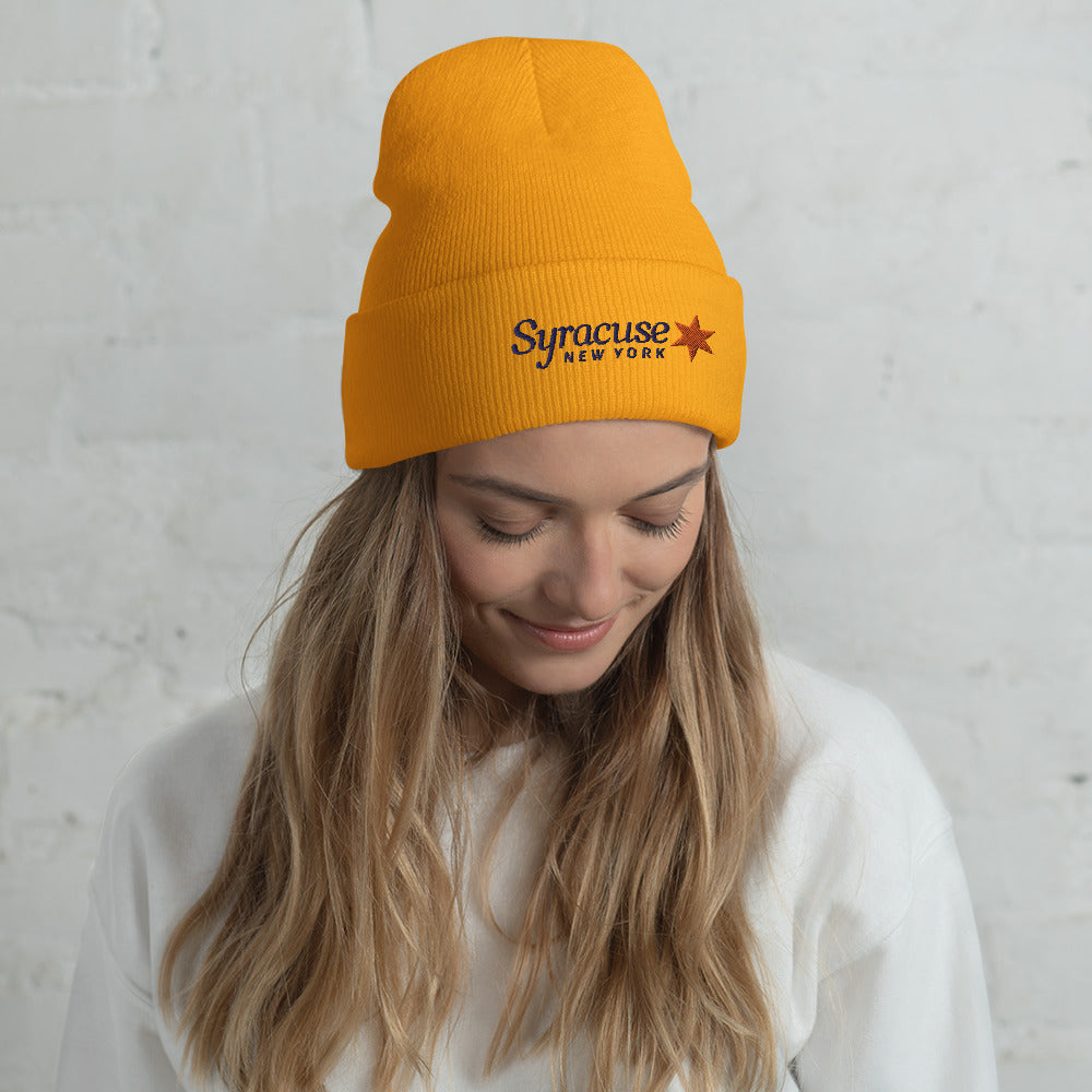 A young woman facing us wearing a yellow-gold winter beanie hat embroidered with a Syracuse, NY logo