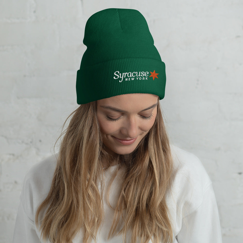 A young woman facing us wearing a spruce green winter beanie hat embroidered with a Syracuse, NY logo
