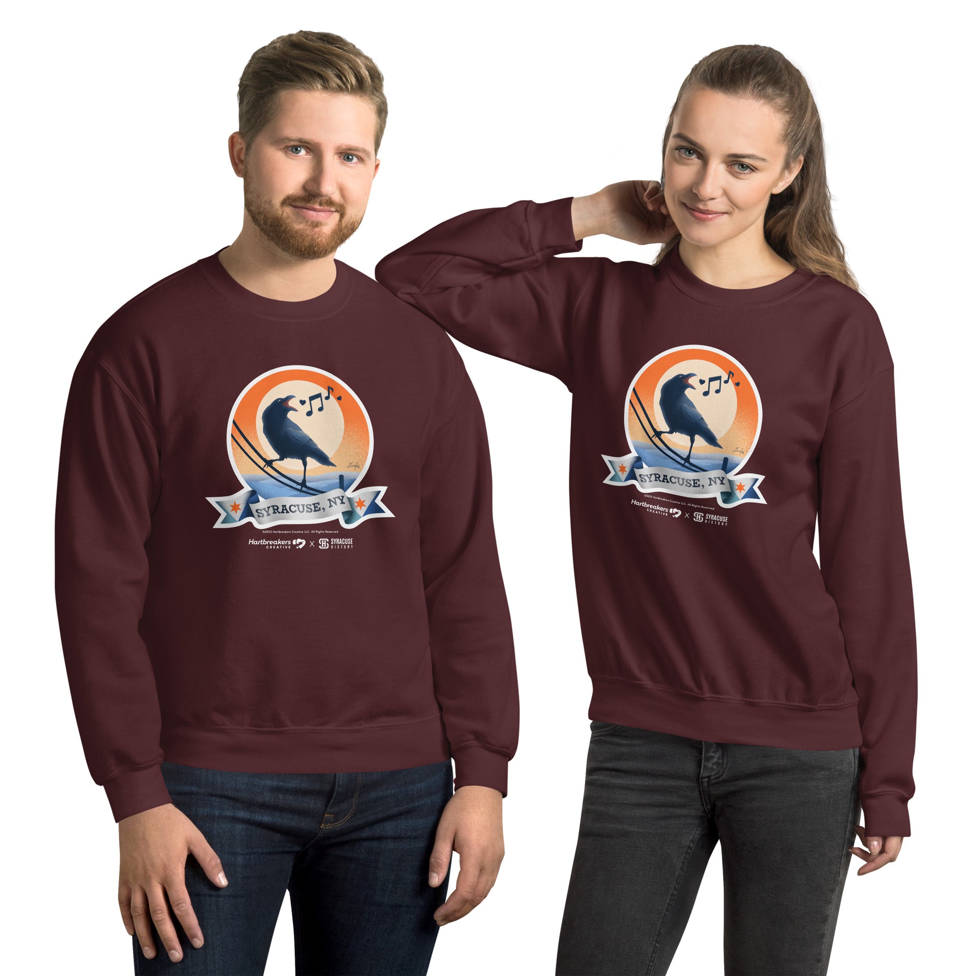 A man and woman wearing maroon sweatshirts featuring an illustration of a crow on a telephone wire and a banner beneath it that says Syracuse, NY