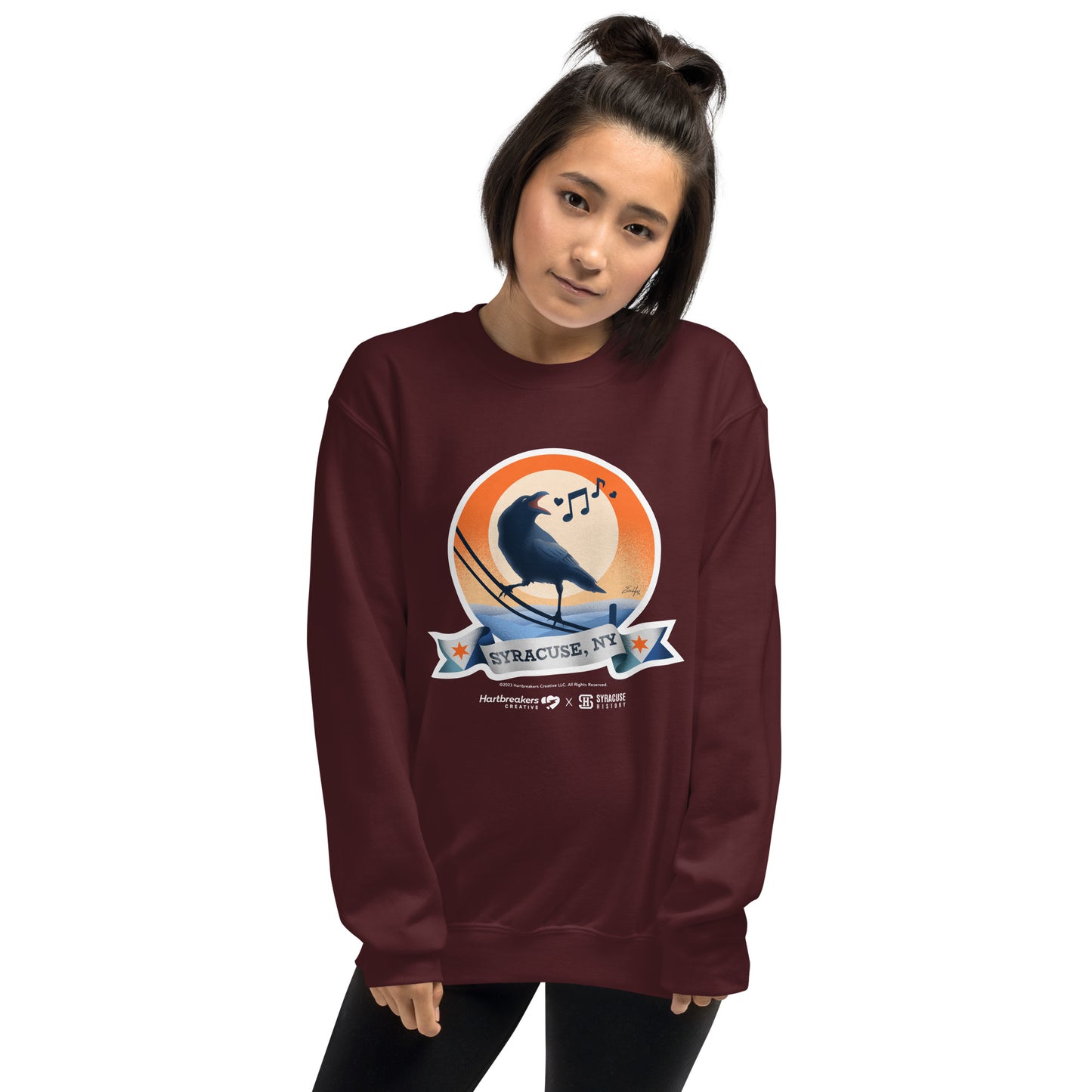 A woman wearing a maroon sweatshirt featuring an illustration of a crow on a telephone wire and a banner beneath it that says Syracuse, NY