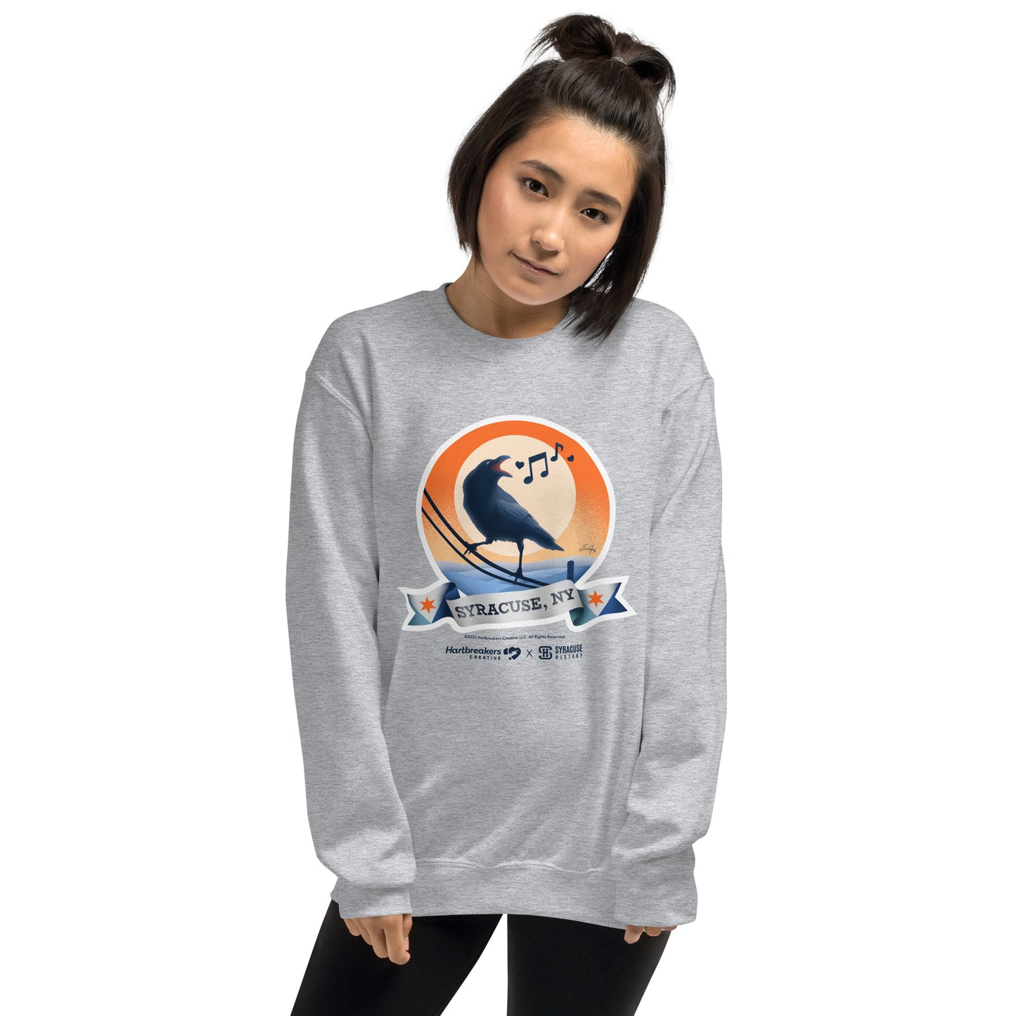 A woman wearing a sport grey sweatshirt featuring an illustration of a crow on a telephone wire and a banner beneath it that says Syracuse, NY