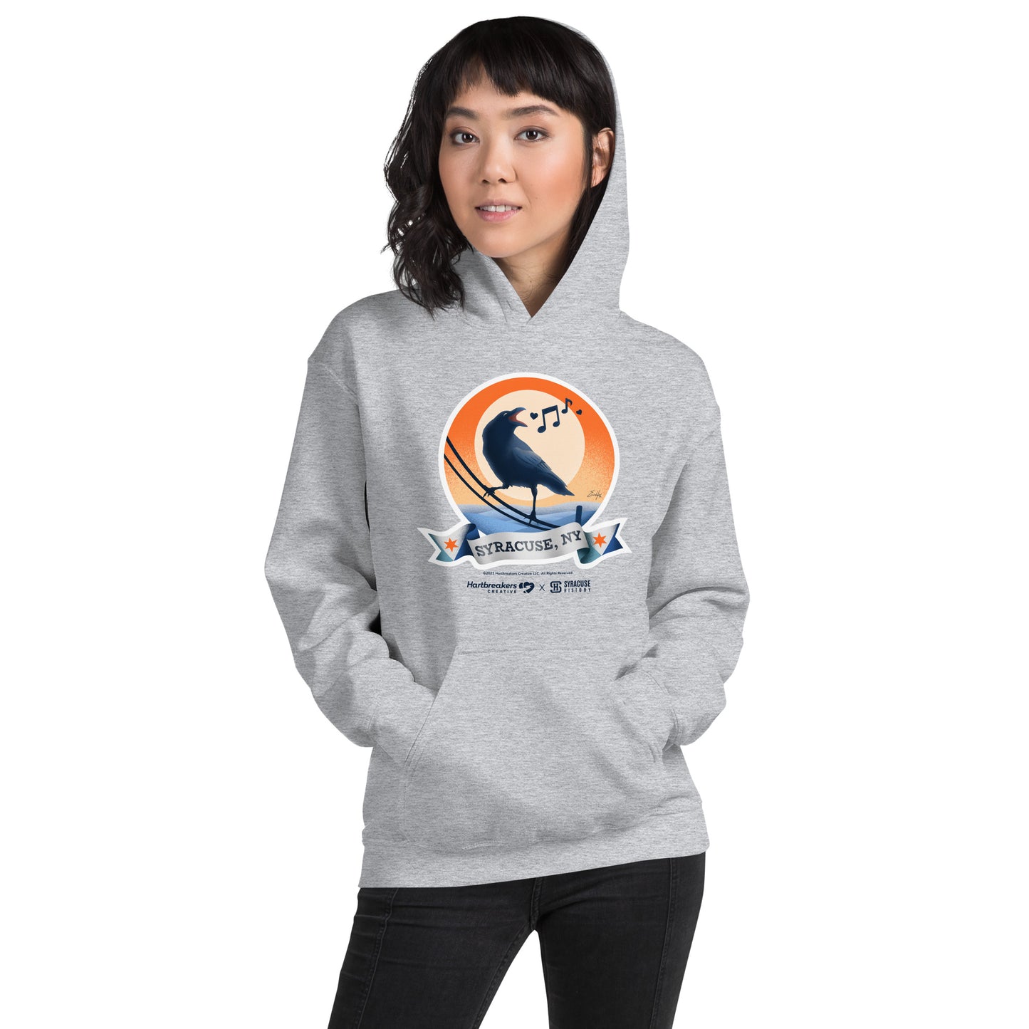 A woman wearing a sport grey hoodie featuring an illustration of a crow on a telephone wire and a banner beneath it that says Syracuse, NY